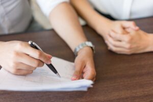 Imagem de duas pessoas com as mãos na mesa e uma delas segurando um contrato com caneta nas mãos, simbolizando os contratos abusivos e práticas comerciais desleais.